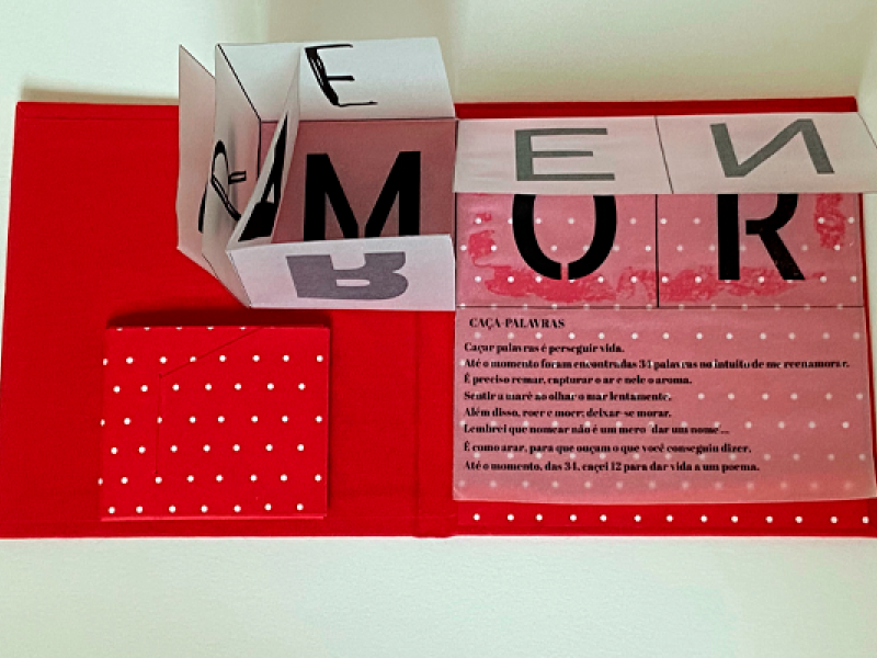 A imagem colorida divulga a exposição “LivroPoema / PoemaLivro”. Em destaque, há um trabalho artístico em vermelho com dobraduras de papel formando letras grandes em preto que compõem palavras.