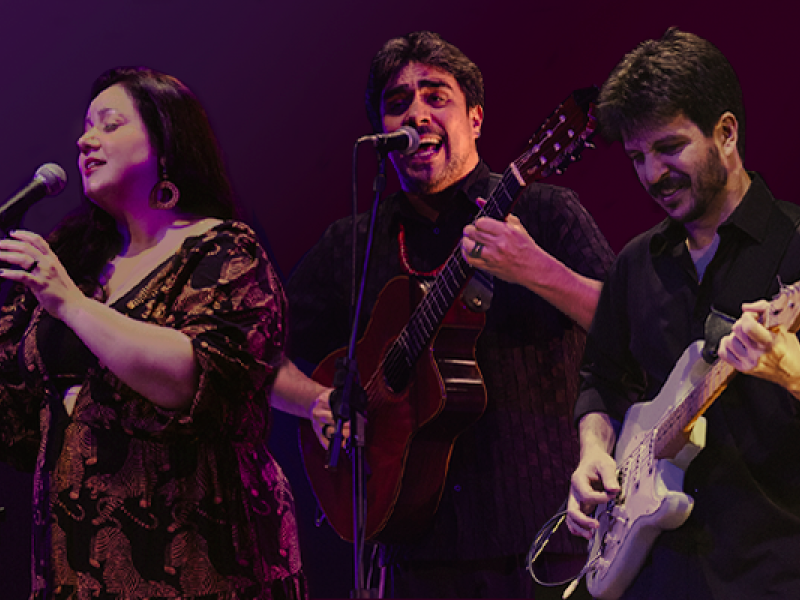 A imagem mostra os músicos Luiza Borges (voz), Thiago Thiago de Mello (voz e violão) e Diogo Sili (guitarras e arranjos) em apresentação. Luiza canta com expressão serena, Thiago interpreta com intensidade ao violão e Diogo toca guitarra com concentração. O fundo em tons de roxo e vermelho valoriza a cena, destacando a energia e harmonia do trio no palco.