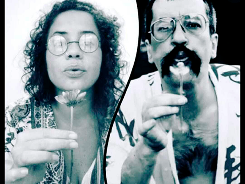 A imagem em preto e branco mostra Nilane Soares Costa e Paulo Leminski, ambos segurando uma flor próxima ao rosto e assoprando suas pétalas. Usando óculos redondos, encaram a câmera de forma expressiva, em um gesto poético que transmite delicadeza e a efemeridade do instante.