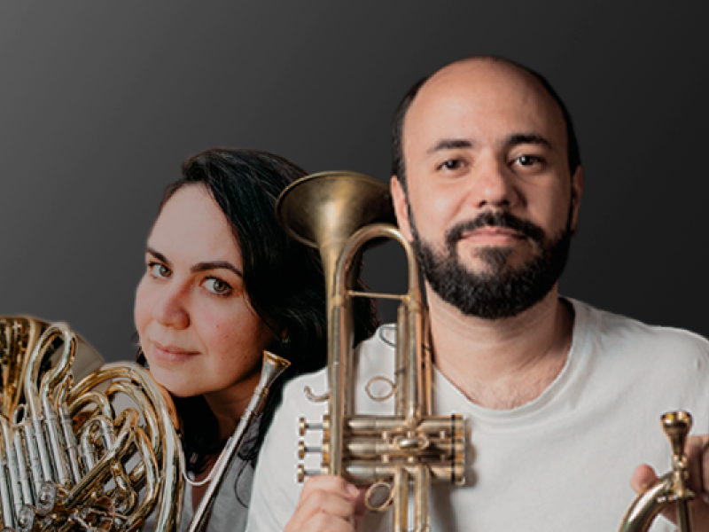 Na imagem, os músicos do Trítono Duo posam com seus instrumentos. À esquerda, Waleska Beltrami segura sua trompa, olhando para a câmera. À direita, Maico Lopes, de barba e camiseta clara, segura um trompete dourado. O fundo em degradê cinza escuro valoriza a dupla e o brilho metálico dos instrumentos, destacando a parceria artística entre os dois.