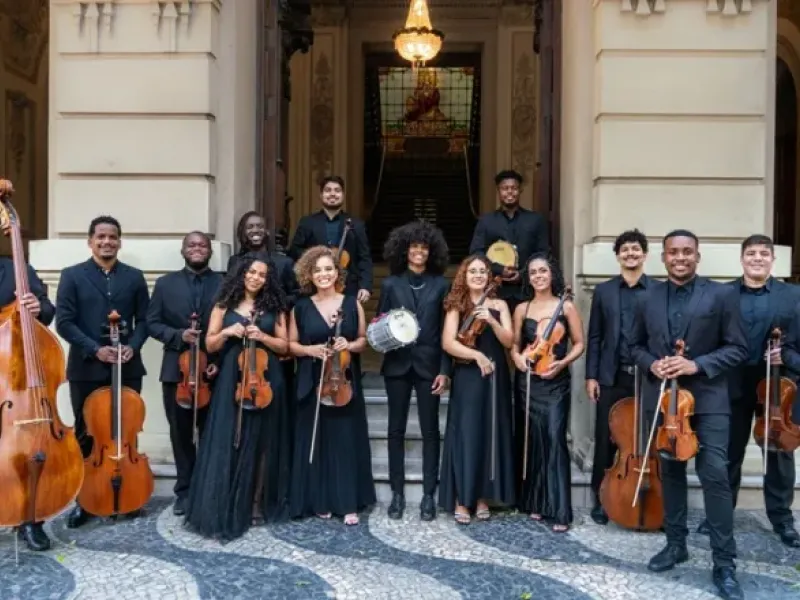 Integrantes da Camerata Jovem do Rio de Janeiro posam sorridentes em frente ao prédio histórico do Centro Cultural Justiça Federal (CCJF). O grupo, formado por 14 músicos vestidos com trajes formais pretos, segura instrumentos como violinos, violoncelos, contrabaixo, pandeiros e triângulo. A escadaria e o vitral iluminado ao fundo compõem o cenário elegante e acolhedor da foto.