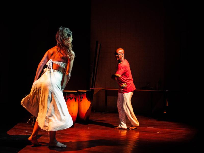 Cena em um palco iluminado por luz âmbar mostra a pianista Verónica Fernandes e o percussionista Anderson Vilmar em um momento de dança e expressão corporal. Verónica, de saia e top brancos, movimenta-se em diálogo com Anderson, que veste camiseta vermelha e calça branca. Ao fundo, um tambor reforça a atmosfera rítmica e ancestral da cena, marcada por energia, conexão e celebração da cultura afro-brasileira.