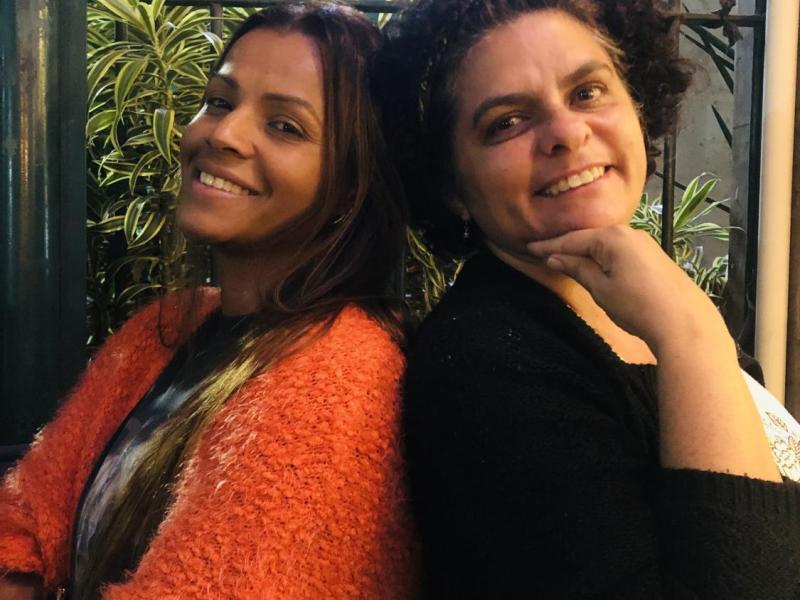As escritoras Luciana Belém e Ednéia Rodrigues posam sorridentes, encostadas uma na outra, diante de um fundo com plantas. Luciana, à esquerda, veste um casaco alaranjado e tem cabelos longos; Ednéia, à direita, usa um casaco preto e apoia o queixo na mão, com expressão acolhedora. A imagem transmite leveza e cumplicidade entre as autoras.