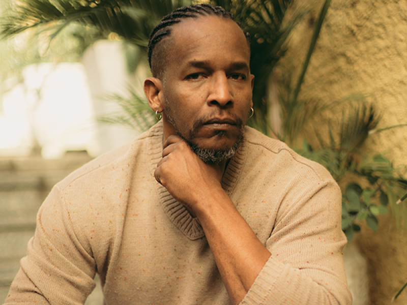 O cantor Alcides Sodré aparece sentado em um ambiente externo, cercado por folhagens. Ele olha diretamente para a câmera com expressão serena e introspectiva, apoiando o queixo sobre a mão. Usa uma blusa de tricô bege, e a iluminação suave valoriza seus traços e o clima de contemplação da cena.