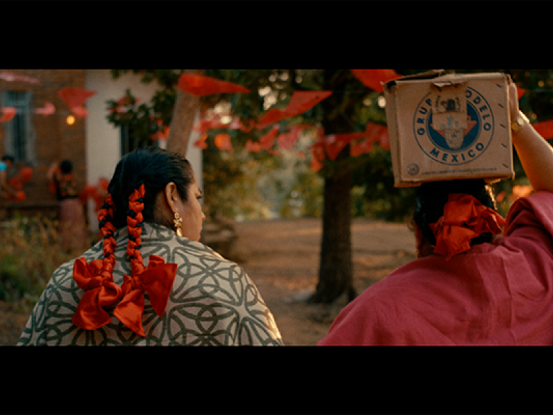 Cena do filme La Baláhna. Duas pessoas caminham por um espaço ao ar livre enfeitado com bandeirolas vermelhas entre as árvores. Vistas de costas, uma usa uma manta estampada e tranças adornadas com grandes laços vermelhos; a outra veste tecido vermelho e equilibra na cabeça uma caixa com o selo “Producto de México”. A luz dourada e o cenário evocam um clima de tradição e celebração popular.