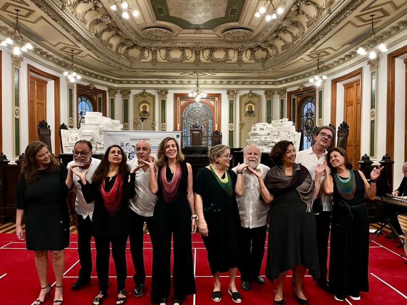 A imagem mostra o Grupo Vocal BateBoca posando de forma descontraída na Sala de Sessões do CCJF, espaço de arquitetura histórica com teto ornamentado, vitrais e detalhes clássicos. Os integrantes, sorridentes, estão sobre um tapete vermelho, vestidos em tons de preto e branco, e alguns usam echarpes coloridas. A cena transmite alegria e sintonia, refletindo o clima festivo após uma apresentação musical.