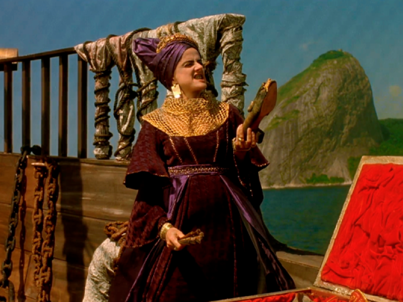 Cena do filme Carlota Joaquina, Princesa do Brazil, em que a atriz Marieta Severo interpreta a personagem principal. Ela aparece em um navio, vestida com traje luxuoso em tons de roxo e dourado, usando toucado e colares ornamentados. À sua frente, há um baú aberto, e ao fundo, o mar e o Pão de Açúcar compõem o cenário histórico da cena.