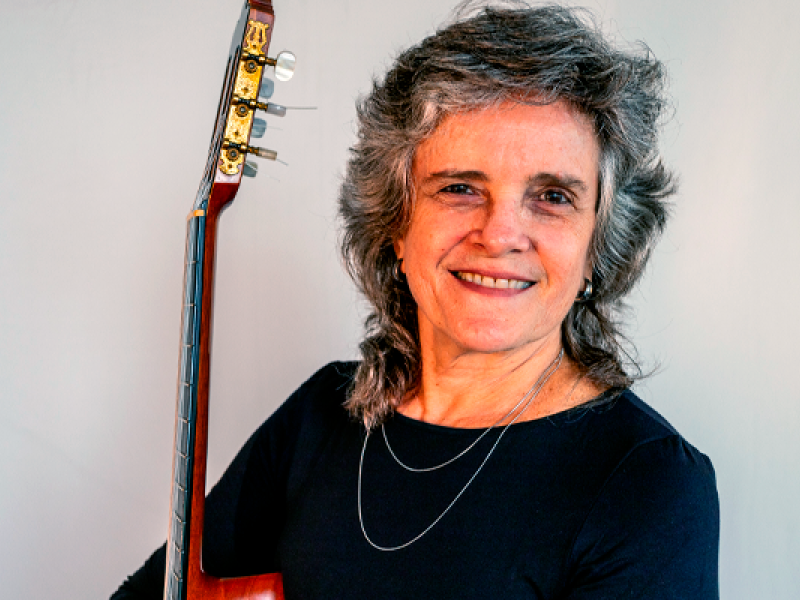 A violonista Vera de Andrade, de cabelos grisalhos e ondulados, sorri suavemente enquanto segura o violão apoiado sobre o ombro. Ela veste uma blusa escura e um colar delicado de duas voltas. O fundo neutro realça sua expressão serena e confiante, refletindo sua conexão com o instrumento e sua trajetória musical.