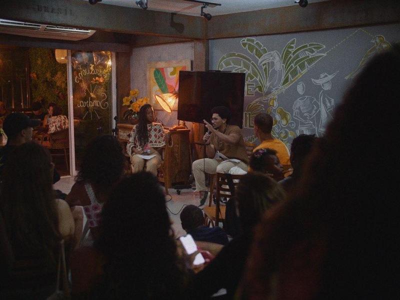 A imagem mostra um espaço cultural intimista, com pessoas sentadas assistindo a uma roda de conversa ou apresentação. À frente, duas pessoas estão no palco: uma mulher de cabelos longos e um homem falando ao microfone. O ambiente é decorado com murais artísticos na parede, iluminação aconchegante e plantas, criando uma atmosfera acolhedora e descontraída.