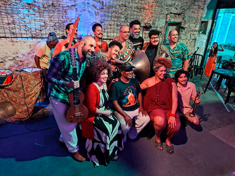Integrantes da Orquestra Balogun posam juntos em um ambiente rústico, com paredes de pedra e iluminação colorida em tons de azul e vermelho. O grupo reúne músicos e artistas com instrumentos como violão, atabaque e outros tambores, formando uma composição vibrante. Todos sorriem e se aproximam para a foto, transmitindo união, celebração e a força da musicalidade afro-brasileira que marca o trabalho da orquestra.