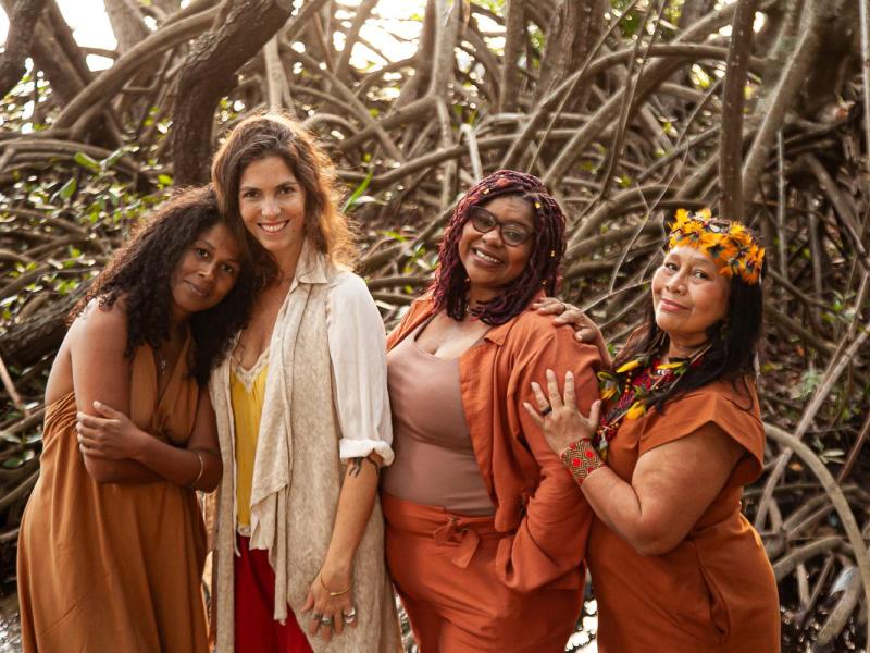 A equipe responsável pelo Raízes – Festival de Arte Sustentável posa unida diante de um cenário natural formado por raízes e galhos entrelaçados. As quatro mulheres, vestidas em tons terrosos que dialogam com a proposta ecológica do festival, sorriem com acolhimento e cumplicidade. A composição transmite conexão com a natureza, ancestralidade e o espírito colaborativo que move o projeto.