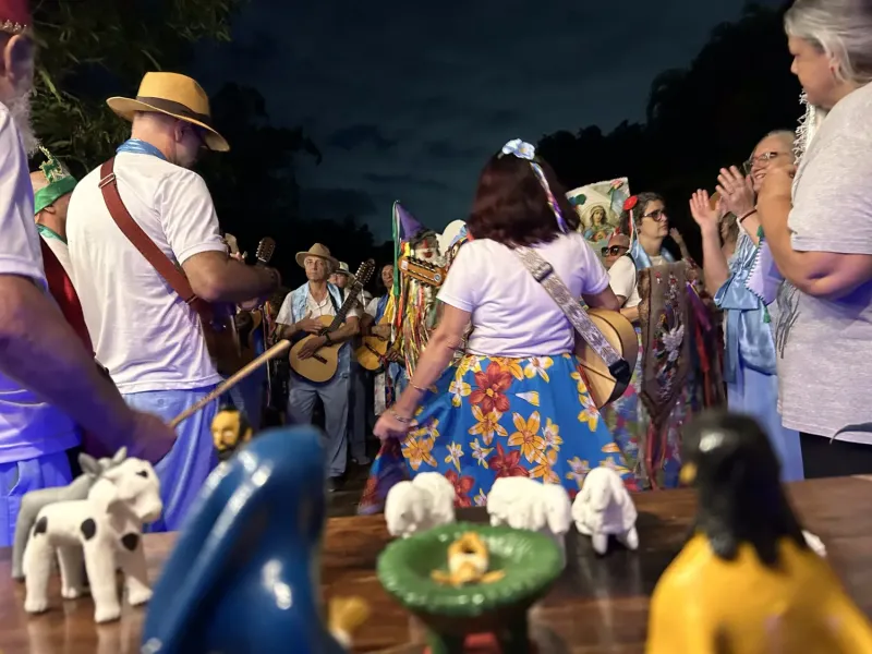 Integrantes de Folia de Reis do Sertão Carioca se reúnem ao ar livre à noite, vestidos com roupas coloridas, chapéus e instrumentos musicais como violas e pandeiros. Em primeiro plano, um presépio artesanal com figuras de barro representa o nascimento de Jesus. Ao fundo, o grupo canta e toca em celebração tradicional, marcada pela devoção, alegria e forte expressão da cultura popular.