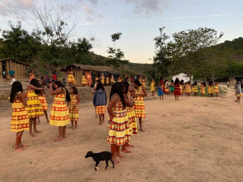 Cena que parece ser de um filme ou documentário, mostrando um grupo de mulheres e meninas indígenas reunidas em uma área aberta de terra batida, diante de casas de madeira. Elas usam vestidos amarelos com faixas vermelhas e se organizam em círculo, enquanto um pequeno cachorro preto aparece em primeiro plano. O entardecer cria uma luz suave sobre o cenário, cercado por árvores e morros ao fundo.