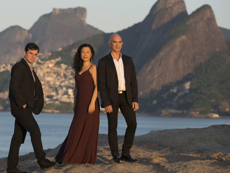 O trio musical T’RIO posa sobre uma formação rochosa à beira-mar, com o Morro Dois Irmãos e o Vidigal ao fundo, banhados pela luz suave do entardecer. Os três integrantes estão elegantes: a mulher veste um longo vestido vinho, enquanto os dois homens usam ternos escuros. A imagem transmite sofisticação e harmonia, refletindo o estilo refinado do grupo.