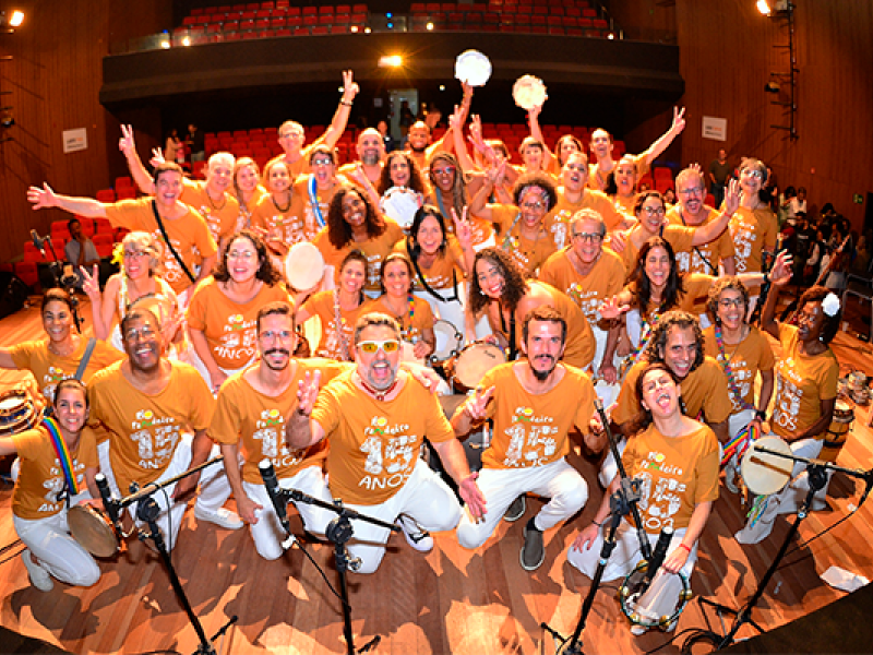 A Companhia Musical Rio Pandeiro reúne-se no palco para uma foto vibrante e cheia de energia. O grande grupo, vestido com camisetas laranja comemorativas, sorri, ergue instrumentos e faz poses animadas. Pandeiros, tambores e outros instrumentos de percussão destacam-se na cena. Ao fundo, veem-se as poltronas vermelhas do teatro, reforçando o clima de celebração e espetáculo coletivo.