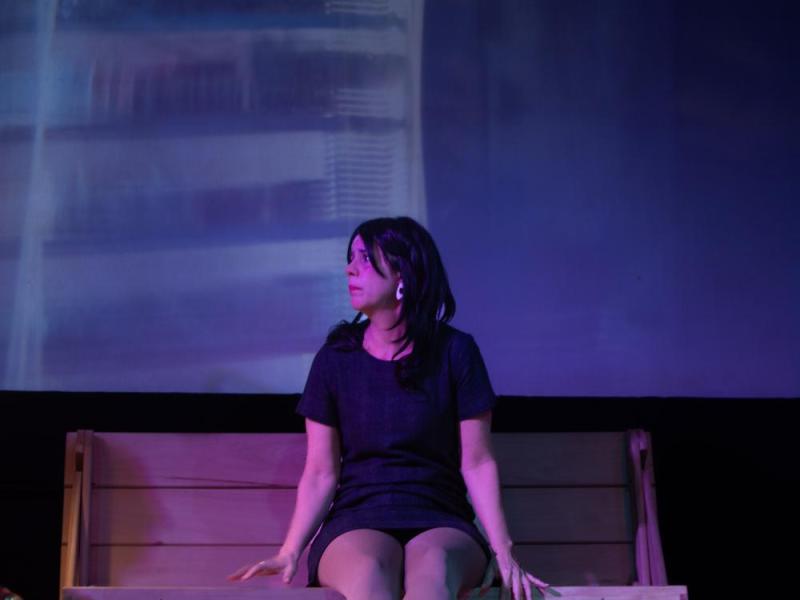 A foto mostra uma mulher sentada em um banco de madeira, em um ambiente de iluminação cênica em tons de roxo e azul. Ela usa um vestido escuro e olha para o lado, com expressão pensativa. Ao fundo, vê-se um painel desfocado que reforça o clima teatral da cena.