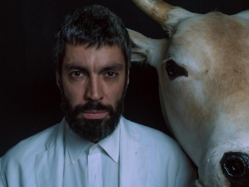 A foto mostra um homem de barba e cabelos curtos posando ao lado da cabeça de um boi branco. Ele usa uma roupa clara e olha diretamente para a câmera, com expressão séria. O fundo escuro destaca as duas figuras em primeiro plano, criando um contraste forte entre o rosto humano e o animal.