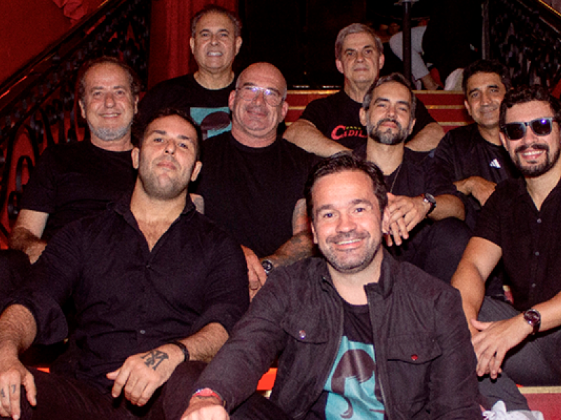 Grupo de nove homens, integrantes da banda de Rock ’n’ Roll Cadillac, está reunido em uma escada de ambiente interno, posando para a foto. Todos vestem roupas escuras e sorriem para a câmera. A iluminação quente destaca os rostos e o corrimão ornamentado ao fundo, criando uma atmosfera descontraída e vibrante, típica do universo do rock.