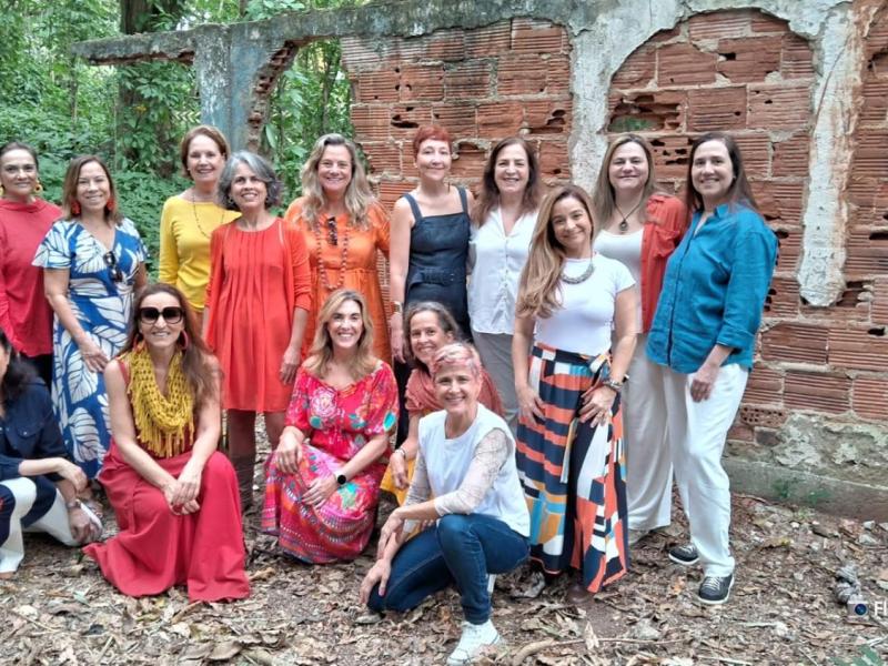 Integrantes do Acorde Vocal, grupo vocal feminino, posam juntas ao ar livre diante de uma antiga parede de tijolos aparentes. As mulheres vestem roupas coloridas e variadas, algumas em pé e outras agachadas, sorrindo para a câmera. O cenário natural, com folhas e vegetação ao redor, transmite acolhimento, união e a força coletiva do grupo.