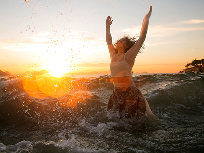 Mulher está no mar, com a água chegando à cintura, de braços erguidos em gesto de celebração. Ela usa blusa clara e saia estampada, enquanto ondas se quebram ao redor. Ao fundo, o sol se põe no horizonte, criando luz dourada e reflexos na água, transmitindo sensação de liberdade, alegria e conexão com a natureza.