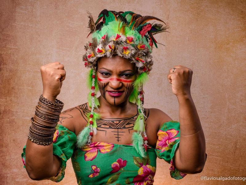 A imagem mostra a cantora Lizza Dias em um retrato de estúdio. Mulher negra, com expressão firme e confiante, olhando diretamente para a câmera. Ela usa um vestido verde com estampa floral e um cocar colorido com penas, sementes e fios verdes. O rosto tem pinturas corporais em vermelho e preto. Ambos os braços estão erguidos, com os punhos fechados em gesto de força. O fundo é neutro, em tom terroso.