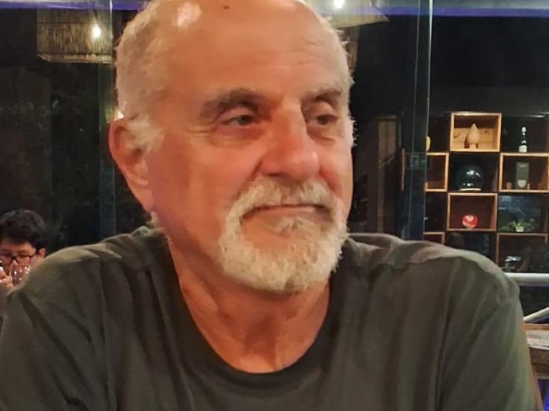 Retrato do professor Alberto Cipiniuk, um homem branco, de aparência madura, com cabelos curtos e grisalhos e barba branca bem aparada. Ele veste camiseta escura e está sentado, com os braços apoiados à frente do corpo. Seu rosto está levemente voltado para a direita, com expressão serena e olhar atento, como se estivesse ouvindo alguém. Ao fundo, o ambiente é interno, com iluminação quente e estantes com objetos decorativos desfocados, sugerindo um espaço cultural ou acadêmico.