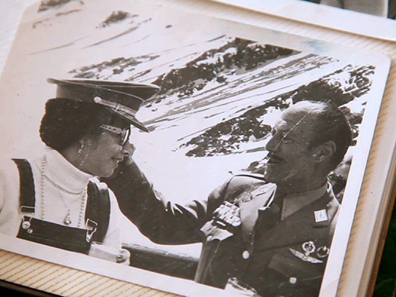Imagem apresentada no documentário O pacto de Adriana. Trata-se de uma fotografia antiga em preto e branco colada em página de álbum, com borda clara e textura levemente amarelada, marcada por discretos riscos diagonais. Na foto, uma jovem de boné militar e jardineira olha para um homem fardado com medalhas, que toca seu rosto em gesto aparentemente afetuoso. Ao fundo, há uma paisagem com água e montanhas. O enquadramento do álbum reforça o caráter de memória e arquivo histórico.