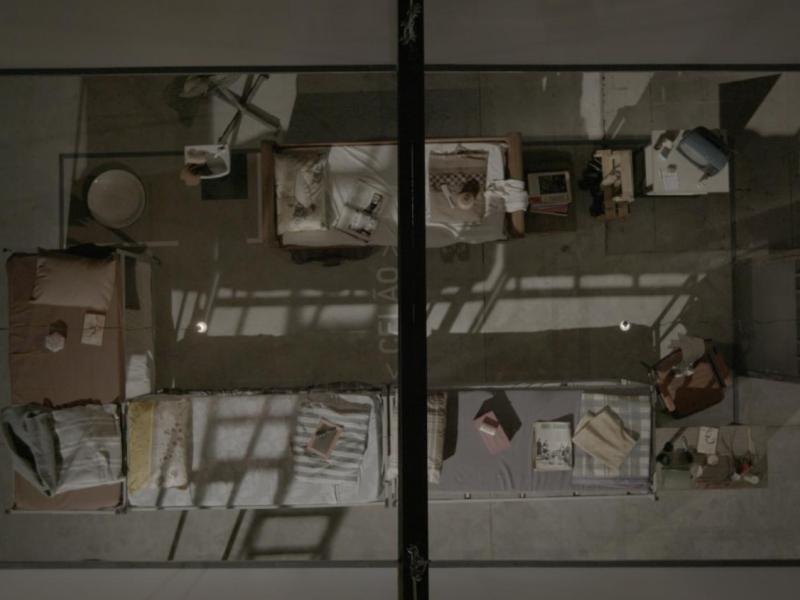 Imagem vista de cima de uma cela coletiva, mostrada como cena do documentário Torre das Donzelas: Memórias e Resistências de Presas Políticas da Ditadura Militar. O espaço é dividido por grades e sombras projetadas, com colchões finos no chão, cobertores dobrados e poucos objetos pessoais espalhados, como livros, roupas e recipientes. A composição rígida e simétrica transmite sensação de confinamento, vigilância e precariedade, reforçando o contexto de repressão e resistência vivido pelas presas políticas.