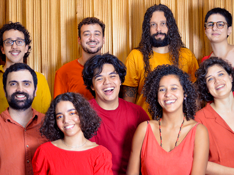 Fotografia do grupo de forró Pena que Voa. A imagem mostra nove pessoas reunidas, posando de forma próxima e descontraída diante de um fundo em tom dourado. O grupo é diverso em gênero, tons de pele e estilos, com cabelos curtos, longos, lisos e cacheados. Todas sorriem ou demonstram expressão alegre e acolhedora. As roupas são leves e coloridas, predominando tons de vermelho, laranja e amarelo, que remetem à energia e à alegria do forró. A composição transmite união, vitalidade e clima festivo.