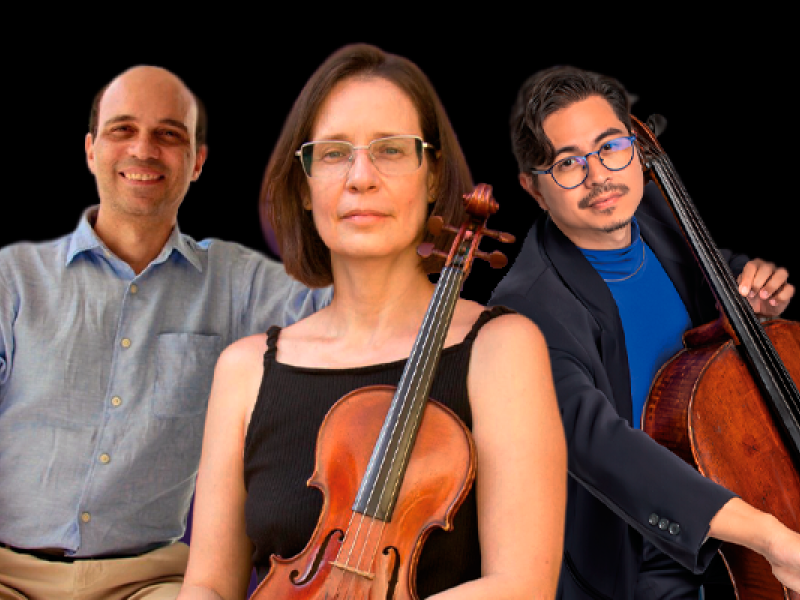 Imagem do Trios de Beethoven e Guerra-Peixe. Três músicos posam diante de fundo preto, em composição próxima e formal. Ao centro, uma mulher segura um violino apoiado junto ao corpo; usa blusa preta sem mangas e óculos, com expressão concentrada e serena. À esquerda, um homem de camisa clara sorri levemente. À direita, outro músico, de óculos e traje escuro com camisa azul, apoia um violoncelo ao lado do corpo. A imagem transmite sobriedade, entrosamento e atmosfera de concerto de música de câmara.