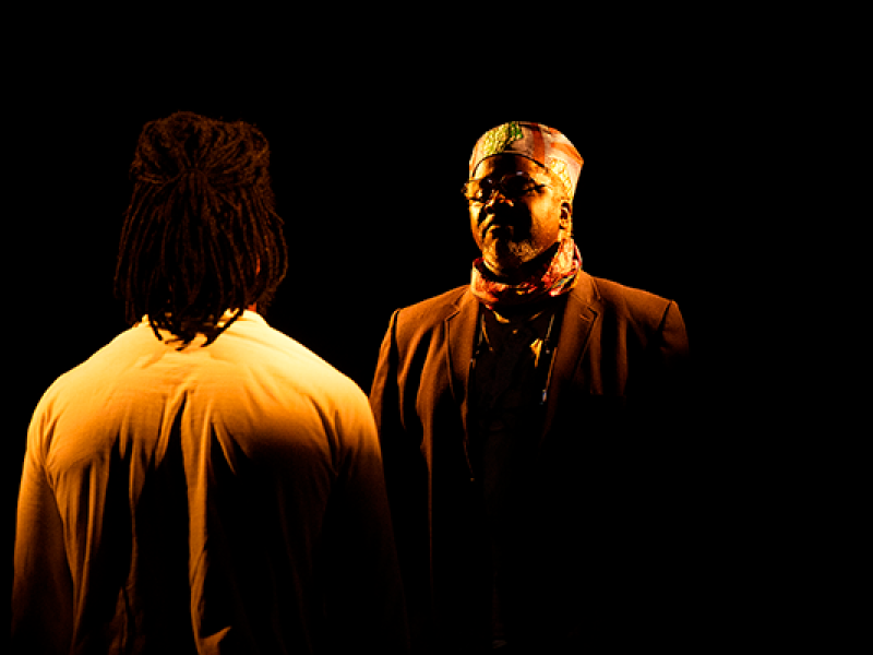 Fotografia de cena teatral da peça “Abdias do Nascimento”. Em palco escuro, dois atores estão frente a frente sob iluminação quente e concentrada. À direita, o ator Lincoln Oliveira aparece de frente, usando terno escuro, óculos, lenço no pescoço e um chapéu de inspiração africana. À esquerda, de costas para a câmera, está o ator Fernando Porto, com cabelos em dreadlocks e camisa clara. O fundo preto destaca o encontro dramático entre os personagens.