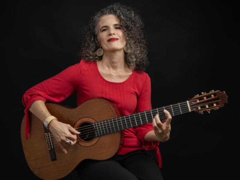 Fotografia de retrato da cantora e compositora Anna Paes. Ela é uma mulher de pele clara, com cabelos cacheados grisalhos, usando blusa vermelha brincos dourados e batom vermelho. Sentada, segura um violão e posiciona os dedos nas cordas, como se estivesse tocando. Olha levemente para cima com expressão serena e confiante. O fundo é preto, destacando a artista e o instrumento.