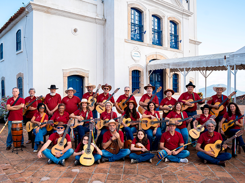 A imagem mostra a Orquestra Caipirando reunida ao ar livre para uma foto coletiva. O grupo é formado por várias pessoas vestidas com camisas vermelhas e chapéus, muitas segurando violas e violões. Alguns integrantes estão sentados no chão ou em bancos, enquanto outros permanecem em pé atrás. Ao fundo, vê-se um edifício histórico de paredes brancas e janelas azuis, com uma área coberta ao lado. O ambiente ensolarado e descontraído destaca o caráter coletivo e musical do grupo.