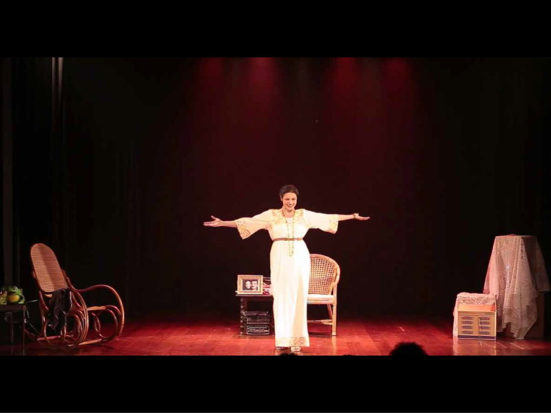 Fotografia de cena teatral com iluminação avermelhada. No centro do palco, a atriz Luciana Borghi está em pé com os braços abertos, interpretando a peça “Na Casa do Rio Vermelho: O Amor de Zélia e Jorge”. Ela veste um vestido longo branco com detalhes dourados. Ao redor, o cenário inclui uma cadeira de balanço, uma poltrona de palha, uma pequena mesa com objetos e caixas, compondo um ambiente doméstico. O palco tem piso de madeira e fundo escuro.