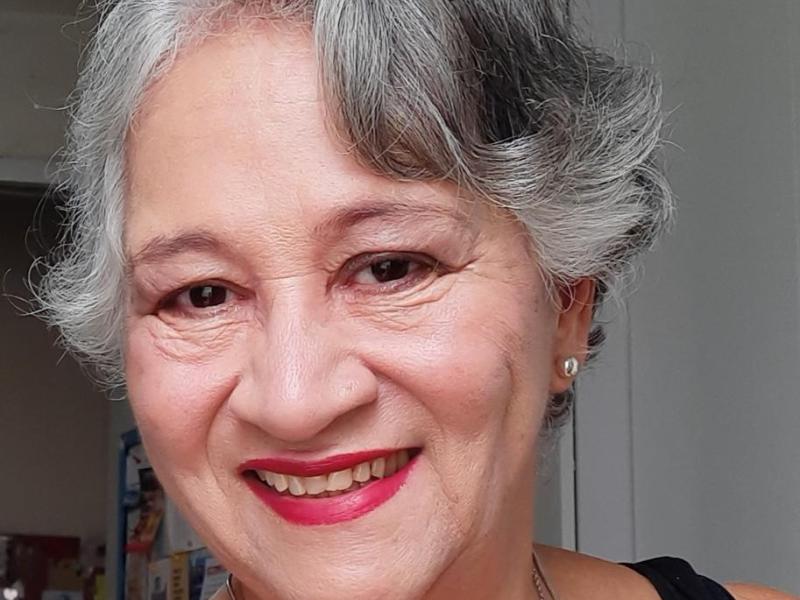 Fotografia de retrato da professora e escritora Denise Gals. Ela é uma mulher de pele clara, com cabelos curtos e grisalhos levemente ondulados. Sorri para a câmera, usando batom vermelho e brincos pequenos. A imagem é um close do rosto, com fundo neutro e desfocado, destacando sua expressão acolhedora.