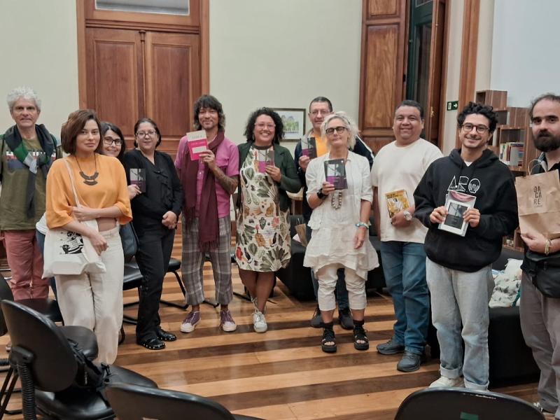 Fotografia de um grupo de cerca de dez pessoas reunidas na sala de leituras do CCJF, durante uma edição anterior do clube de leitura. Os participantes estão lado a lado, sorrindo e olhando para a câmera; alguns seguram livros. O grupo é diverso em idade e aparência. O espaço tem piso de madeira, portas altas de madeira e estantes de livros ao fundo. Em primeiro plano aparecem algumas cadeiras, sugerindo um ambiente de encontro e conversa.