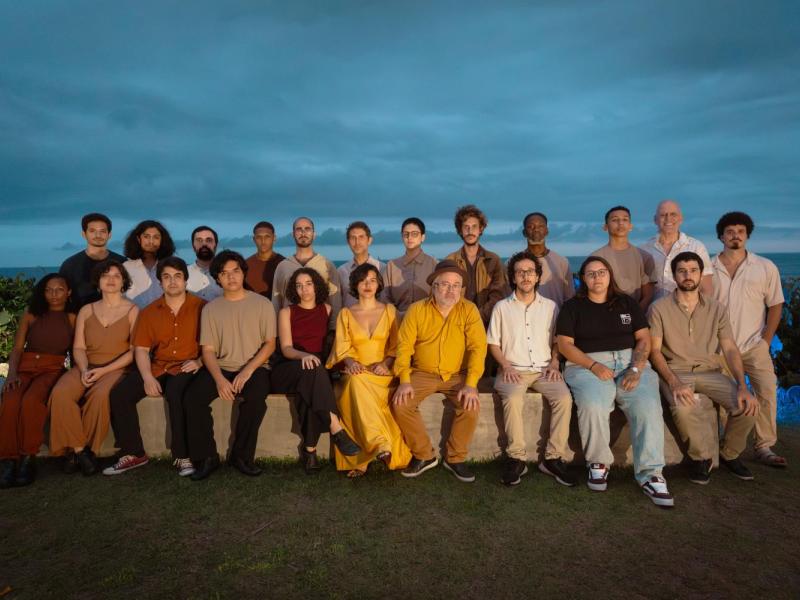 Fotografia de grupo da Orquestra Mil Tons ao ar livre, ao entardecer. Cerca de vinte integrantes estão organizados em duas fileiras — alguns sentados em um banco de pedra e outros em pé atrás. O grupo é diverso em idade, gênero e aparência. Ao fundo, vê-se o mar sob um céu nublado em tons azulados. A iluminação suave destaca os rostos, criando uma atmosfera serena e contemplativa.