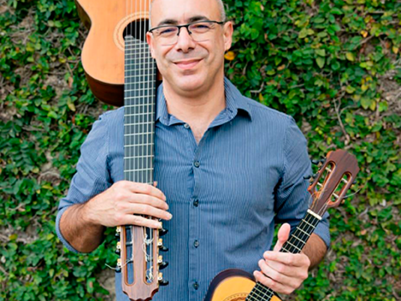 Fotografia de retrato do violonista, cavaquinista, compositor e arranjador Carlos Chaves. Ele é um homem de pele clara, careca, usando óculos e camisa azul de mangas longas. Sorri levemente para a câmera enquanto segura um violão apoiado no ombro e um cavaquinho nas mãos. Ao fundo há uma parede coberta por folhagens verdes, criando um cenário natural que destaca os instrumentos musicais.