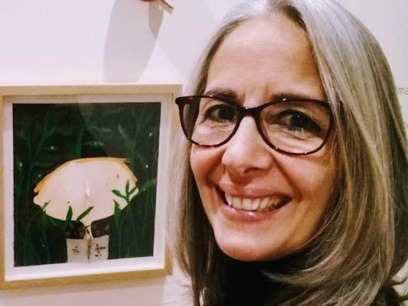 Fotografia de retrato da escritora e artista plástica Patrícia Montês. Ela é uma mulher de pele clara, com cabelos longos e grisalhos, usando óculos de armação escura. Sorri olhando para a câmera. Ao fundo, em uma parede clara, há um quadro emoldurado com uma ilustração de uma luminária cercada por folhas verdes, sugerindo um ambiente de exposição ou galeria.