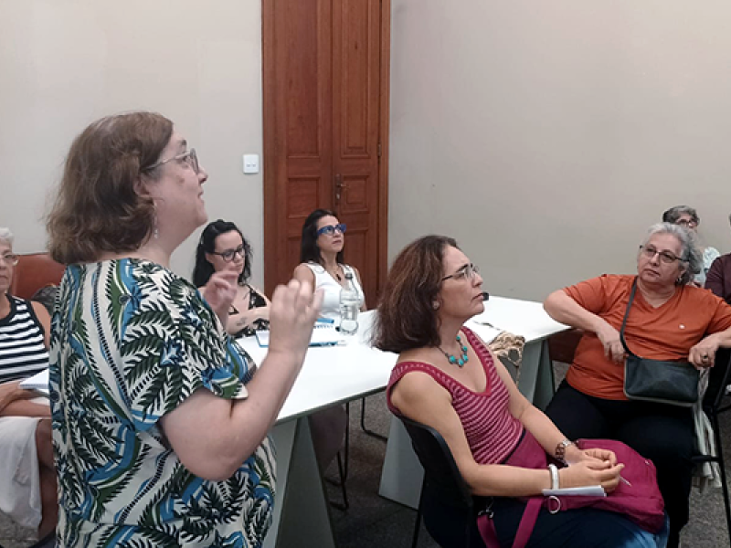 Em uma sala de paredes claras, a escritora e oficineira Cecilia Botana está em pé à esquerda, de óculos e blusa estampada em verde, gesticulando enquanto fala durante uma oficina. À frente, mulheres sentadas ao redor de uma mesa branca escutam com atenção; uma usa vestido rosa e óculos, outra veste blusa laranja e apoia o braço na mesa. Ao fundo há uma porta de madeira e mais participantes acompanhando a atividade.