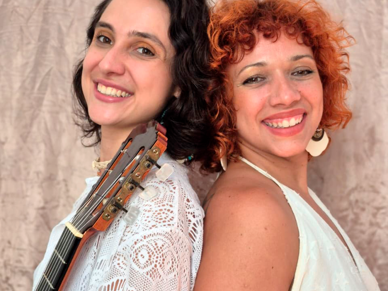 Fotografia de duas mulheres sorrindo, apoiadas costas com costas, diante de um fundo neutro. À esquerda está a violonista Maria Luísa Tonácio, de pele clara e cabelo escuro ondulado, usando blusa branca rendada; o braço de um violão aparece apoiado em seu ombro. À direita está a cantora Deya Mota, de pele clara, cabelos curtos e cacheados em tom ruivo, usando brinco claro e blusa clara sem mangas. Ambas olham para a câmera com expressão alegre.