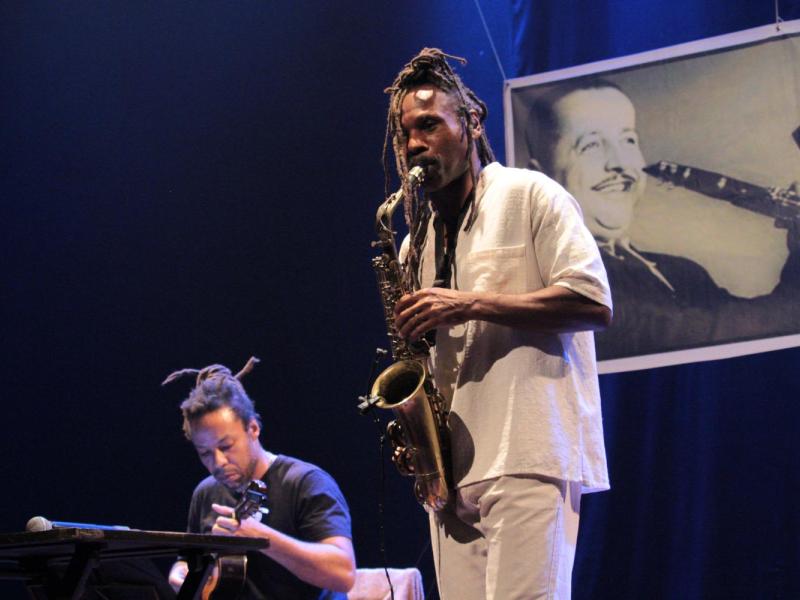 Fotografia de apresentação musical em palco com iluminação azul. Em primeiro plano, o saxofonista Rocino Crispim, homem negro com longos dreadlocks presos no alto da cabeça, toca saxofone vestindo roupa clara. Ao fundo, à esquerda, um dos violonistas da Regional Caboclo aparece sentado, concentrado no violão. Na parte de trás do palco há a projeção de uma foto em preto e branco do músico Luiz Americano tocando saxofone.