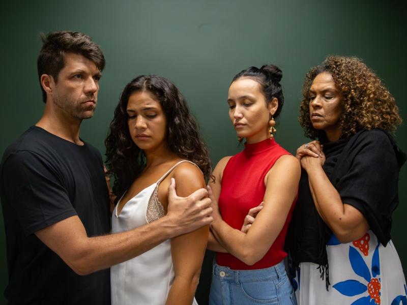 Fotografia da peça teatral O Último Dia. Quatro personagens aparecem lado a lado diante de fundo verde escuro (composição fechada e intensa). À esquerda, um homem segura o braço de uma mulher com expressão séria. As duas mulheres ao centro mantêm os olhos fechados ou baixos, com semblantes tensos e introspectivos. À direita, outra mulher observa com preocupação, mãos unidas junto ao peito. A proximidade dos corpos e as expressões contidas sugerem conflito emocional, luto ou um momento decisivo na narrativa.
