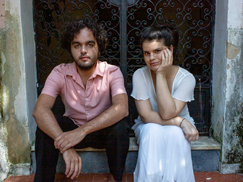 Fotografia de retrato do Duo Bevilacqua Assumpção (violoncelo/piano). A dupla está sentados lado a lado em um degrau, diante de uma porta com grade de ferro trabalhado. À esquerda, o homem de pele clara com cabelos cacheados e barba, veste camisa rosa e calça escura, olhando para a câmera com expressão séria. À direita, a mulher de pele clara com cabelos presos, veste roupa clara e apoia o rosto na mão, sorrindo suavemente. O ambiente tem paredes com marcas do tempo, sugerindo cenário externo ou histórico.