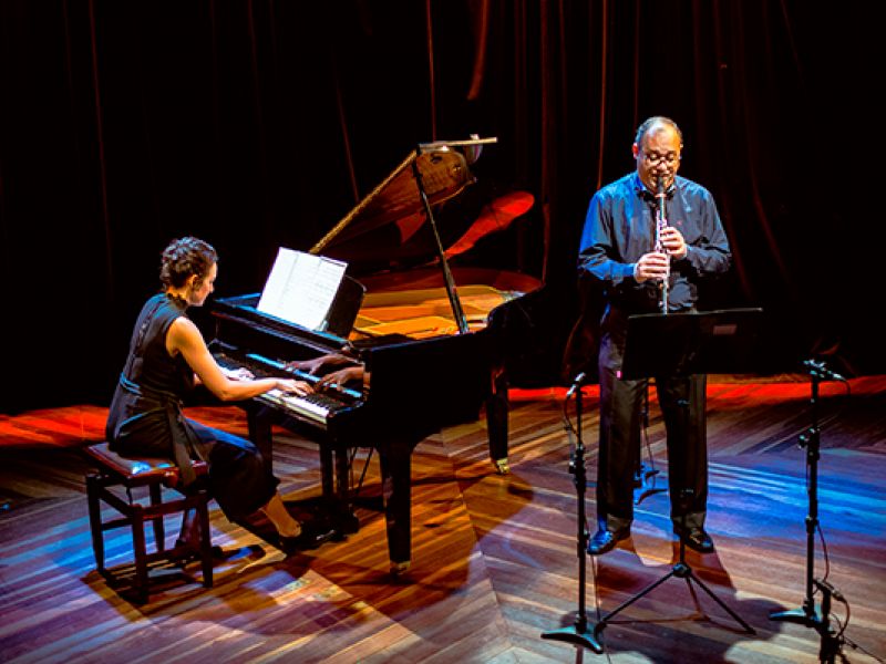 Fotografia de apresentação musical do Duo Ferreira Richter, formado por Bezaleel Ferreira (clarineta) e Raisa Richter (piano). Em um palco com iluminação quente em tons de vermelho e azul, Raisa está sentada ao piano de cauda, lendo a partitura enquanto toca. Ao lado, Bezaleel está em pé, tocando clarineta diante de um suporte de partitura. O chão de madeira e o fundo com cortinas escuras reforçam o ambiente de concerto.