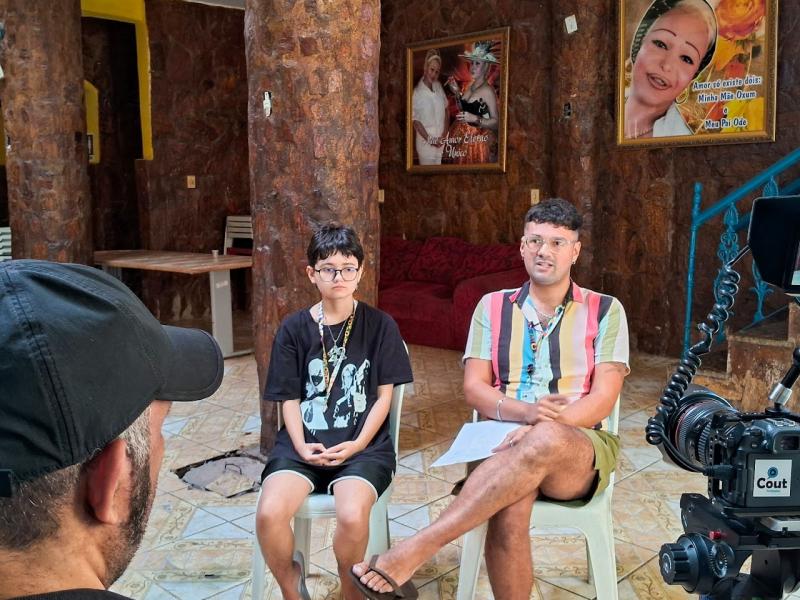 Cena do documentário Dos Armários para as Ruas mostra duas pessoas sentadas lado a lado para uma entrevista. À esquerda, uma criança de óculos, camiseta preta e colar; à direita, um adulto com camisa colorida listrada, short e chinelos, segurando um papel. Em primeiro plano, parte de um cinegrafista e uma câmera profissional indicam a gravação. O ambiente interno tem paredes de pedra, colunas e quadros nas paredes, com clima íntimo e documental.