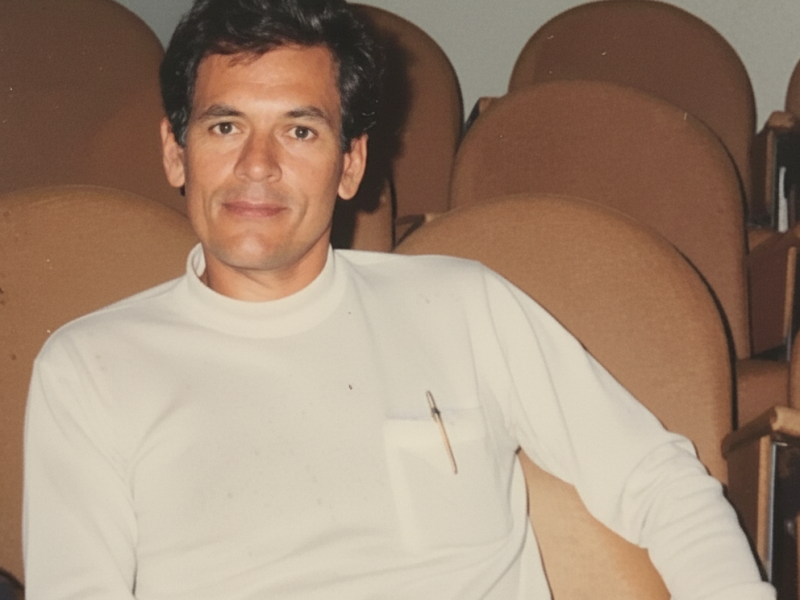 O dramaturgo Roberto Athayde aparece sentado em uma poltrona de auditório, vestindo uma blusa branca de mangas longas com uma caneta no bolso. Ele olha serenamente para a câmera, em meio a fileiras de cadeiras marrons vazias ao fundo, sugerindo um momento de tranquilidade em um ambiente cultural.