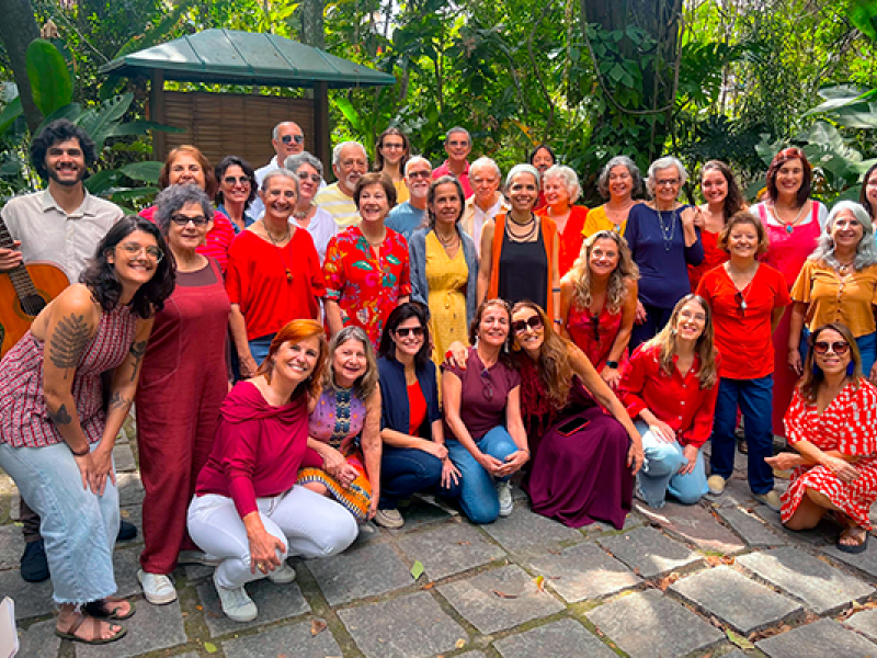 Fotografia de grupo do Coral da Tarde em Canto Plural ao ar livre. Cerca de trinta integrantes estão reunidos em um jardim, entre árvores e plantas, organizados em fileiras — alguns em pé e outros agachados à frente. O grupo é diverso em idade e aparência, vestindo roupas coloridas, com predominância de tons quentes. Todos sorriem e olham para a câmera, transmitindo um clima de alegria e convivência.