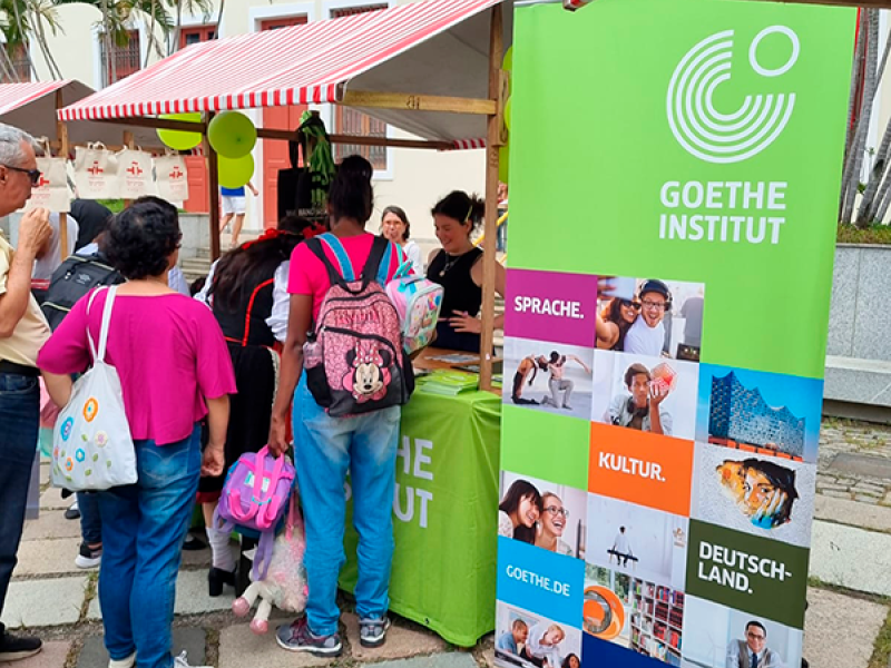 Cena externa parecendo uma feira mostra um estande do Goethe-Institut com cobertura listrada vermelha e branca. Pessoas se aproximam do balcão verde, onde atendentes recebem o público. Uma menina com mochila rosa e um adulto ao lado observam o material. Um banner vertical exibe palavras como “Sprache”, “Kultur” e “Deutschland”, além de imagens ilustrativas. O ambiente é movimentado e de caráter cultural.