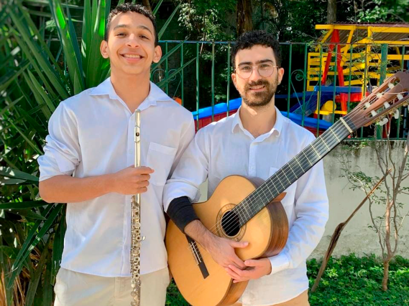 Fotografia de retrato do Duo Sereno, formado pelo flautista Vinícius Marinho e o violonista Iuri Chicharo. Os dois estão lado a lado ao ar livre, sorrindo para a câmera. À esquerda, Vinícius, homem de pele clara, usa camisa branca e segura uma flauta transversal. À direita, Iuri, homem de pele clara com barba e óculos, também veste camisa branca e segura um violão. Ao fundo há vegetação e uma área com brinquedos coloridos, sugerindo um espaço externo ensolarado.